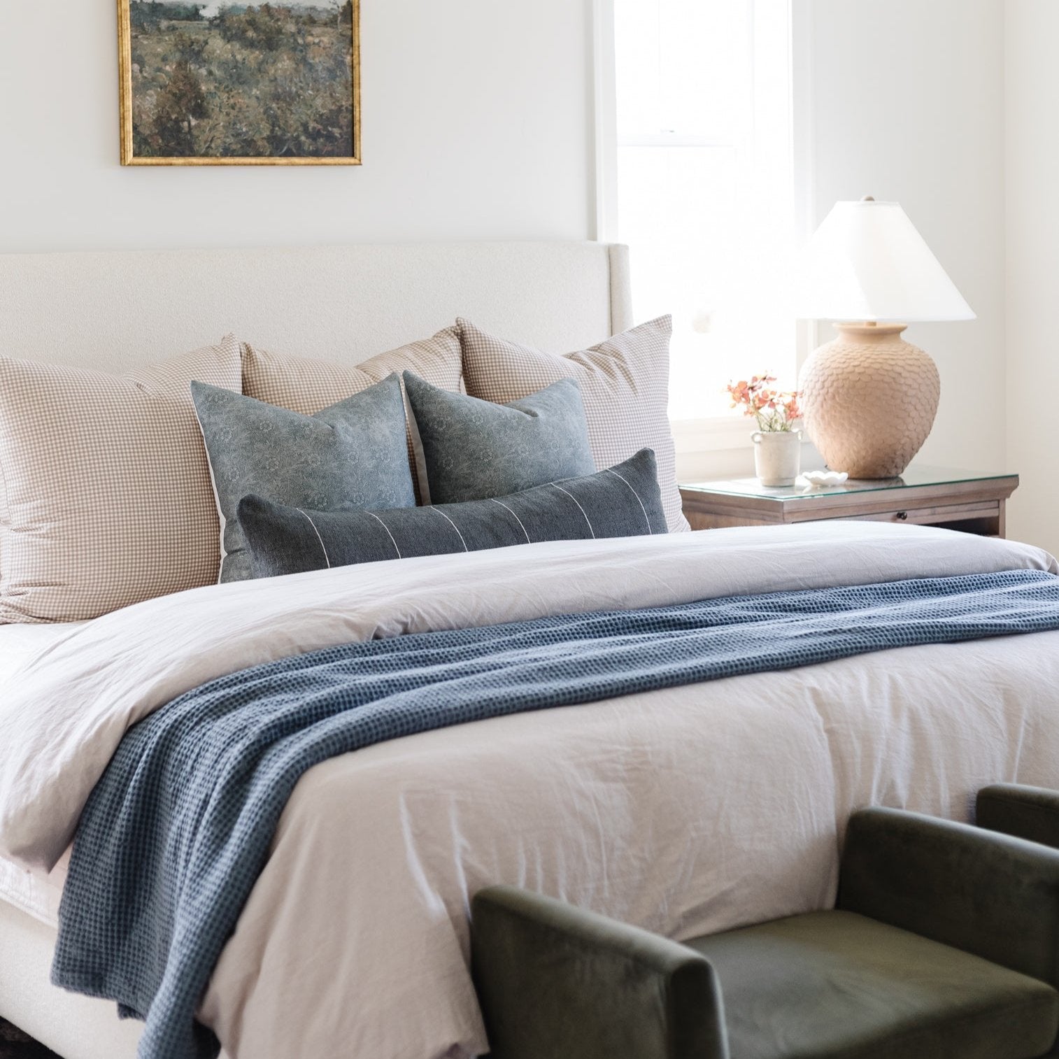 Framed landscape artwork with moss and forest tones displayed above a bedroom bed with blue striped runner and decorative pil