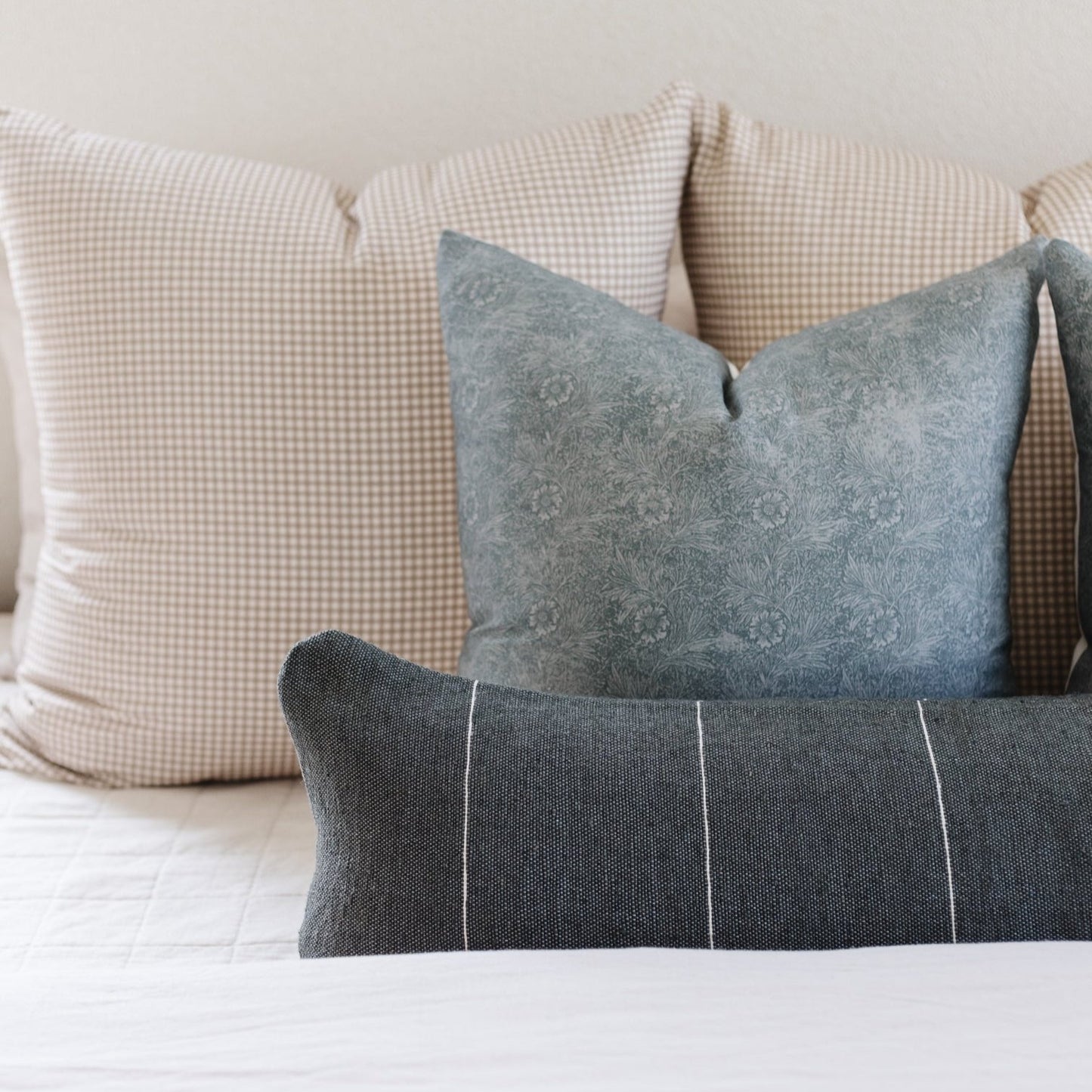 Tan and white gingham check pillow displayed with coordinating gray and patterned pillows on a bed.