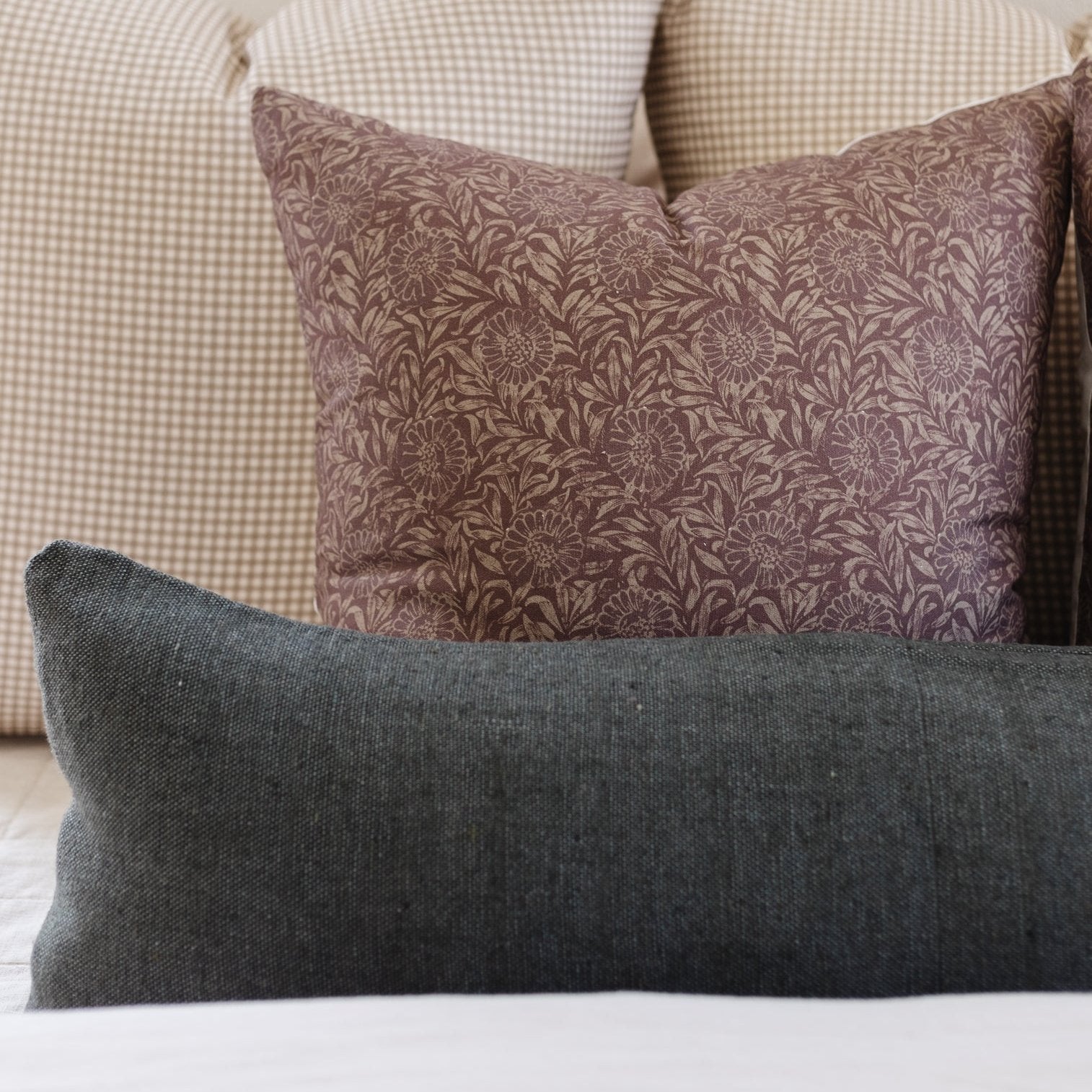 Gray lumbar pillow with charcoal floral patterned square pillow and beige gingham pillows arranged on bed.