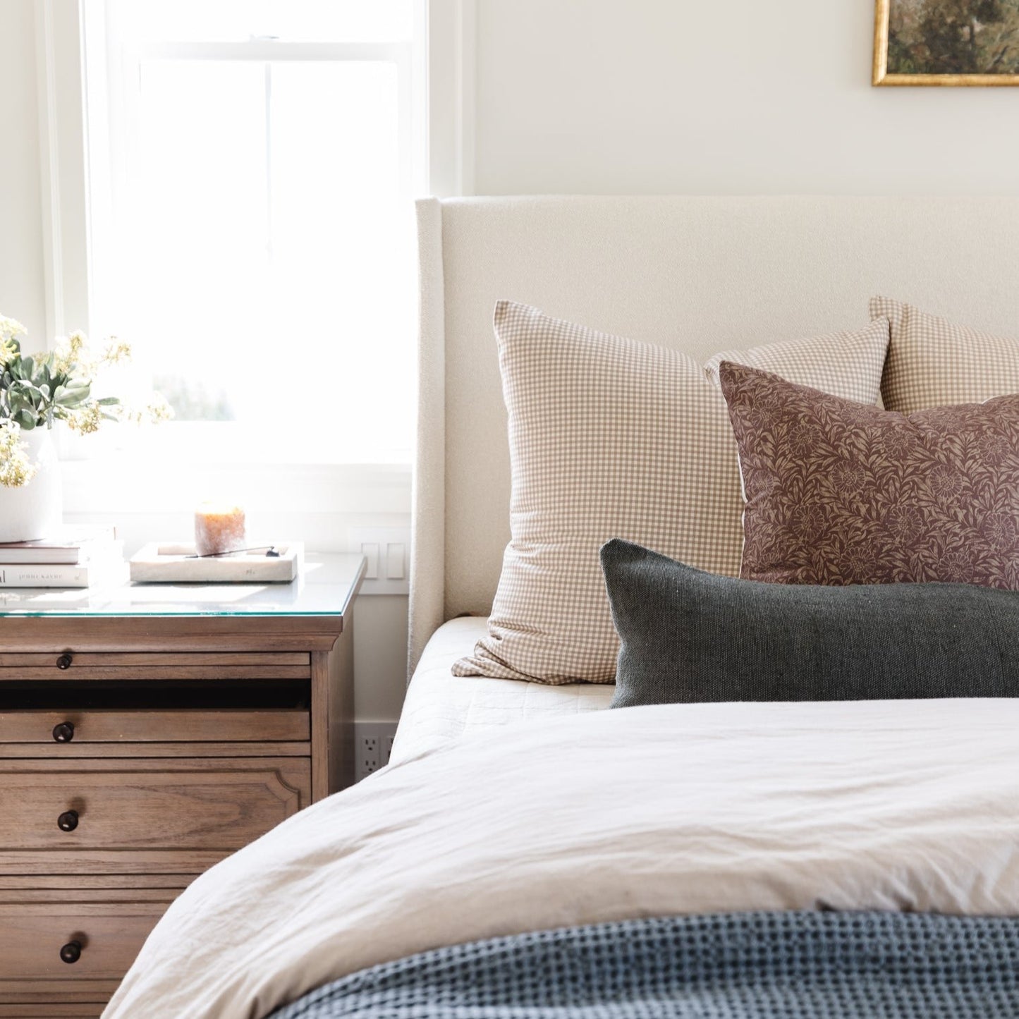 Tan and white gingham patterned pillow on a bed with neutral bedding and wooden nightstand.