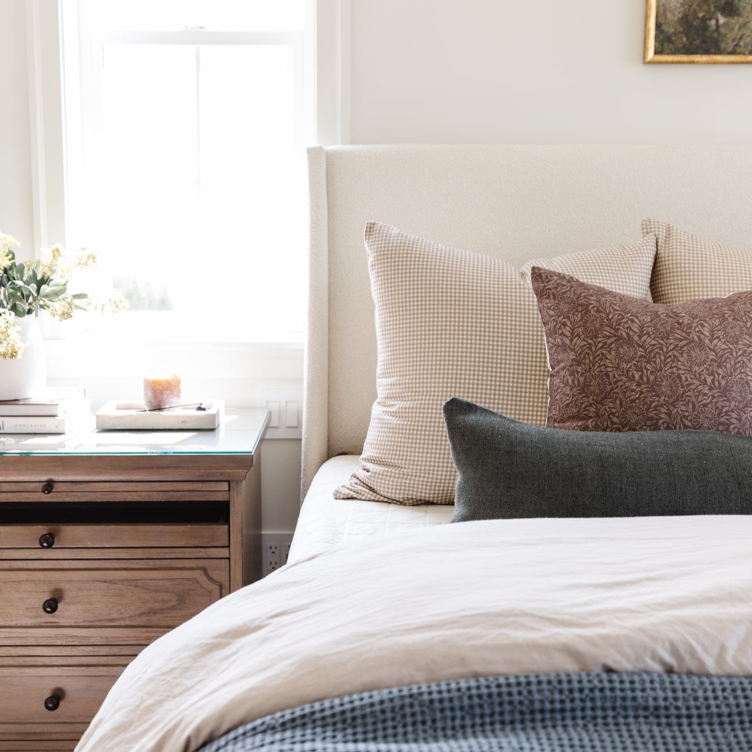 Tan and white gingham patterned pillow on a bed with neutral bedding and wooden nightstand.