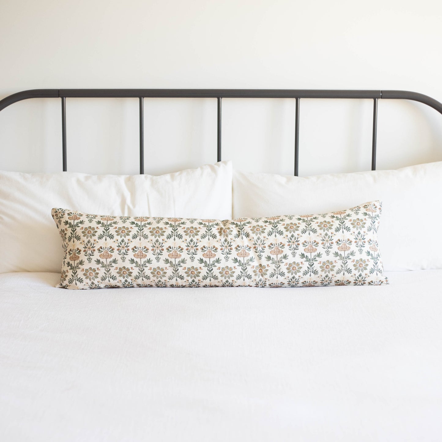 Long rectangular lumbar pillow with floral print in neutral tones on white bedding against metal headboard.