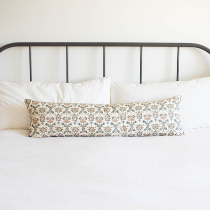 Long rectangular lumbar pillow with floral print in neutral tones on white bedding against metal headboard.