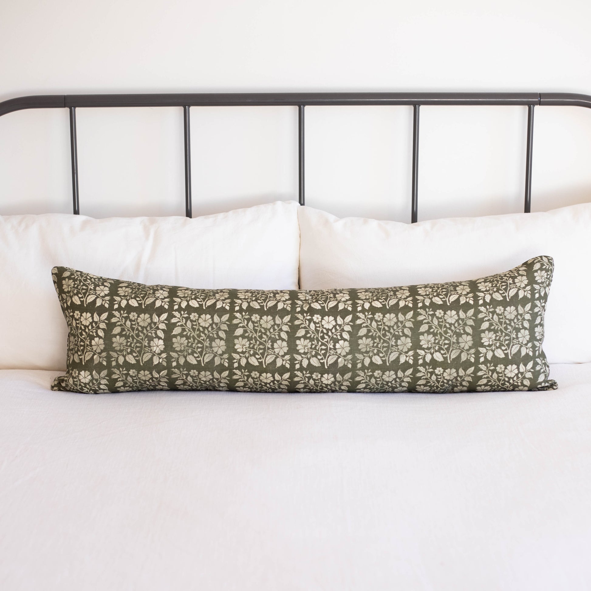 Sage green linen lumbar pillow with white floral leaf pattern on bed with white pillows and metal headboard.