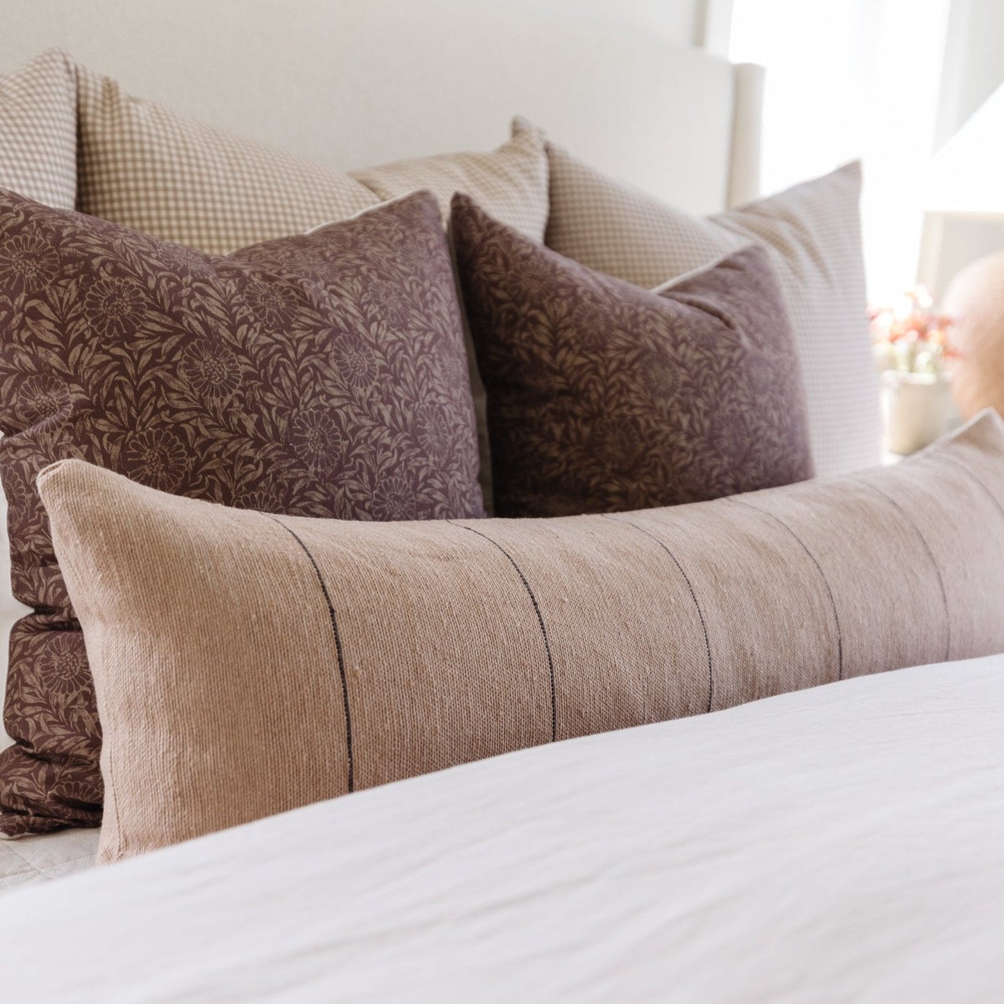 Beige lumbar pillow with pleated texture displayed among patterned brown and checkered pillows on a bed.