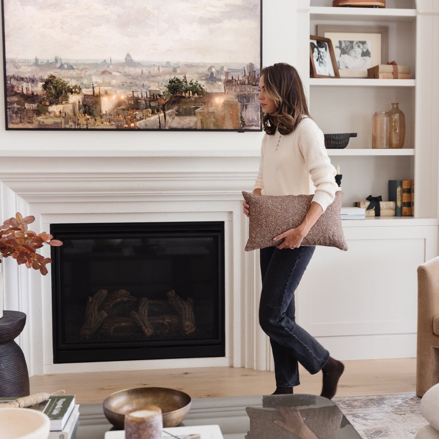 Taupe textured lumbar pillow held by woman in living room with fireplace and framed cityscape art.