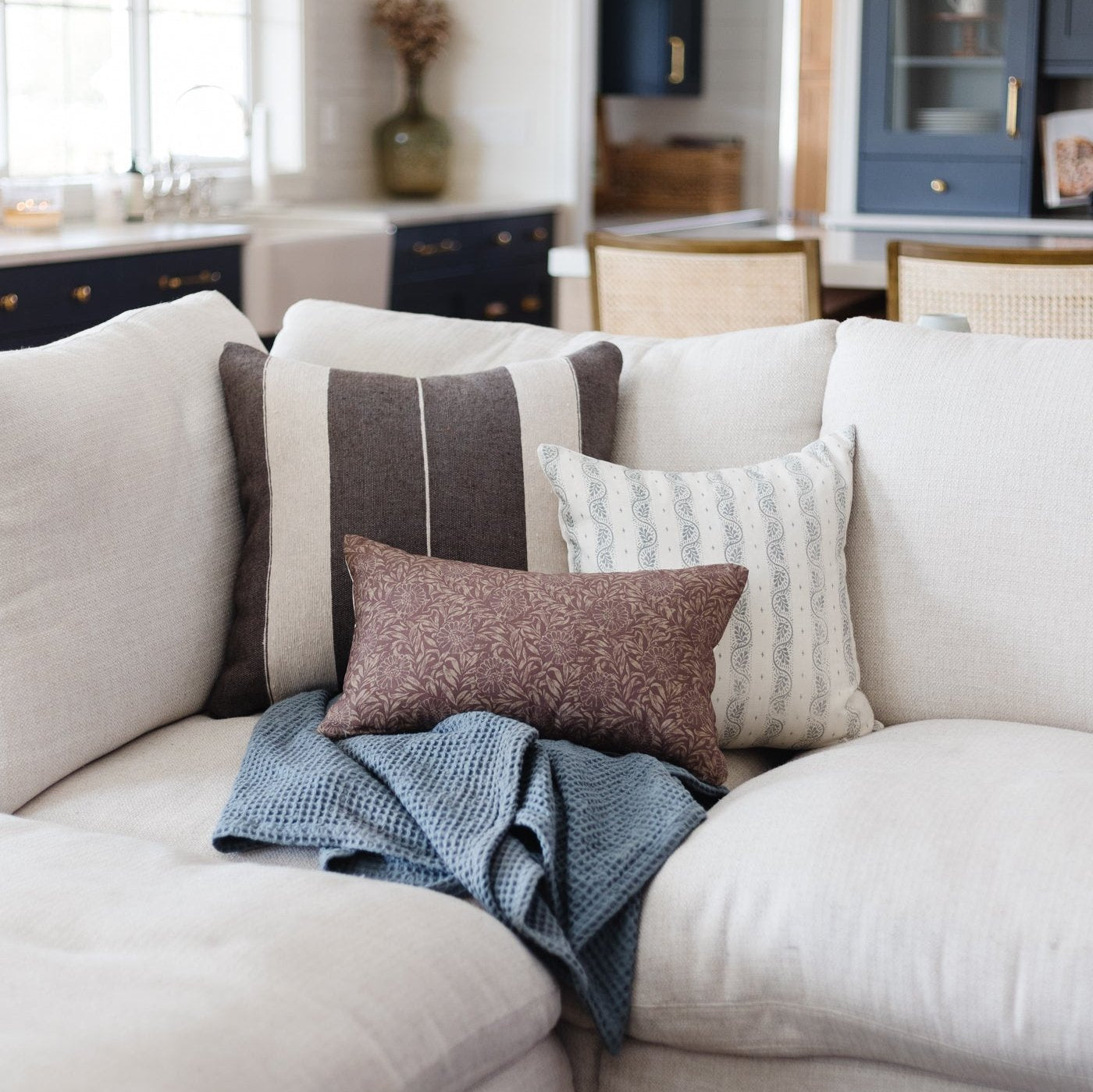 Brown damask lumbar pillow with white piping displayed on a cream sofa with other decorative pillows and a blue throw blanket