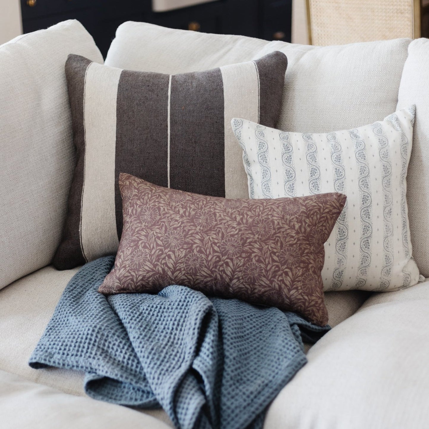 Decorative pillows in cream, gray, and patterned fabrics arranged on a beige sofa with a blue throw blanket.