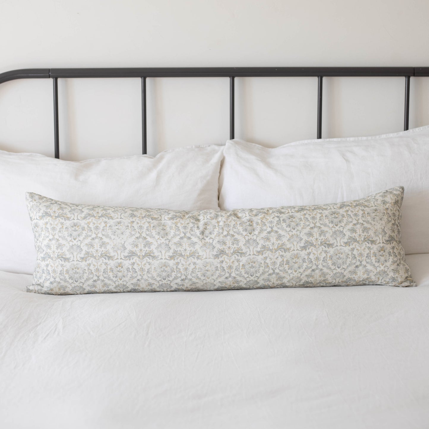 Long rectangular throw pillow with beige floral pattern resting on white bedding against black metal headboard.