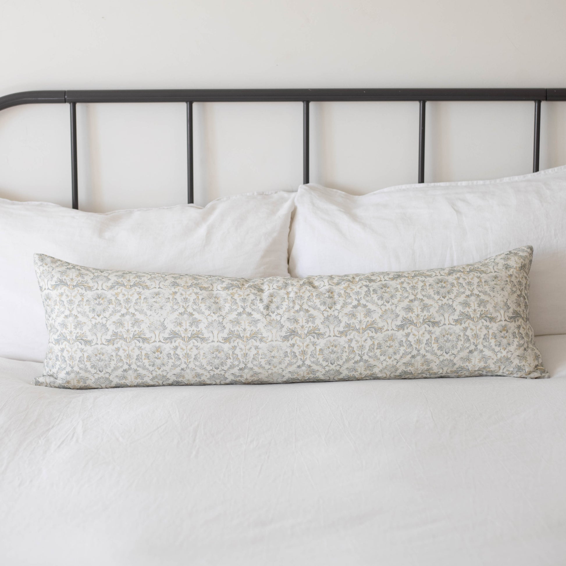Long rectangular throw pillow with beige floral pattern resting on white bedding against black metal headboard.
