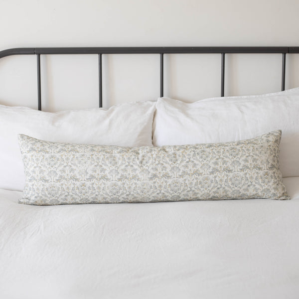 Long rectangular throw pillow with beige floral pattern resting on white bedding against black metal headboard.