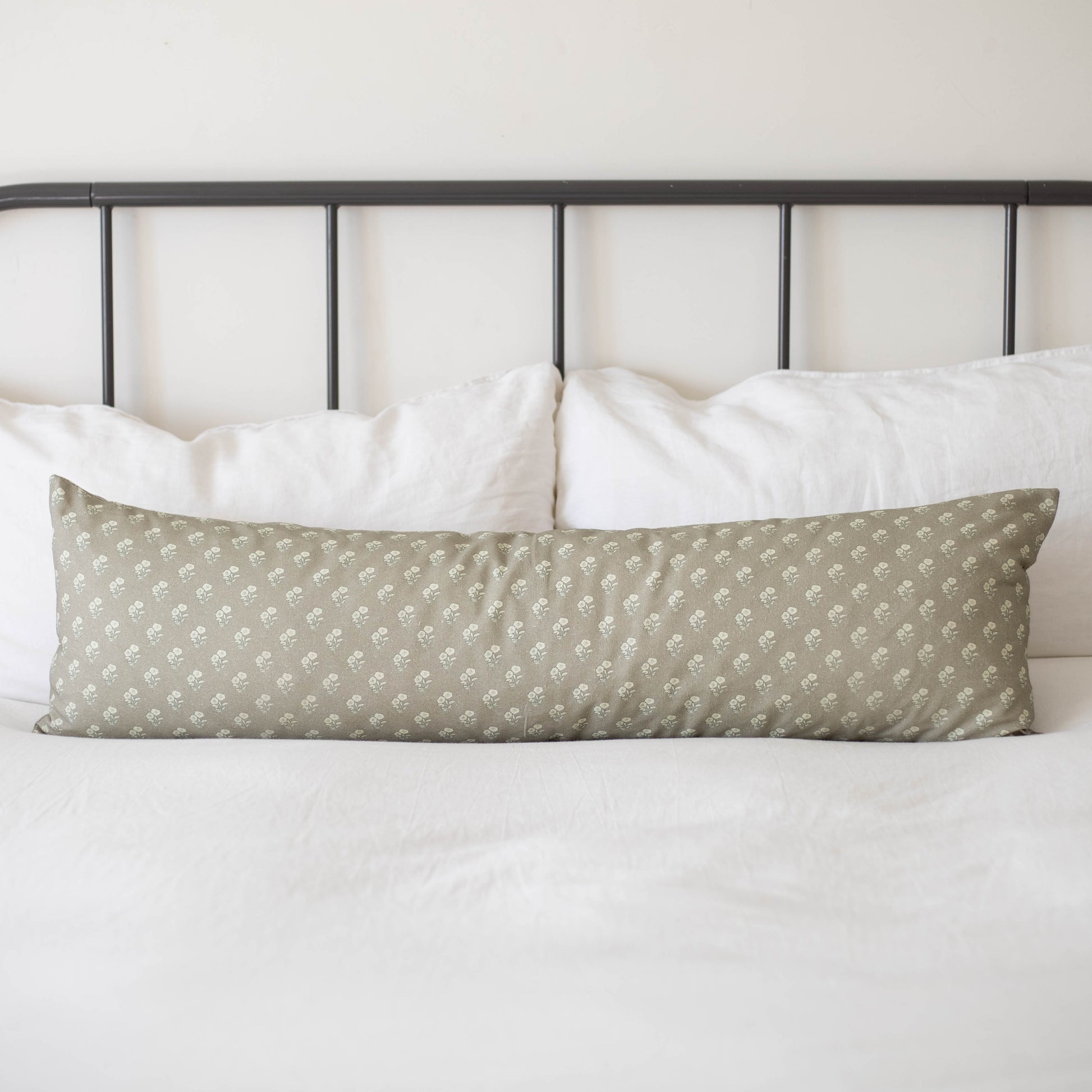 Sage green lumbar pillow with small white floral pattern on bed with white pillows and metal headboard.
