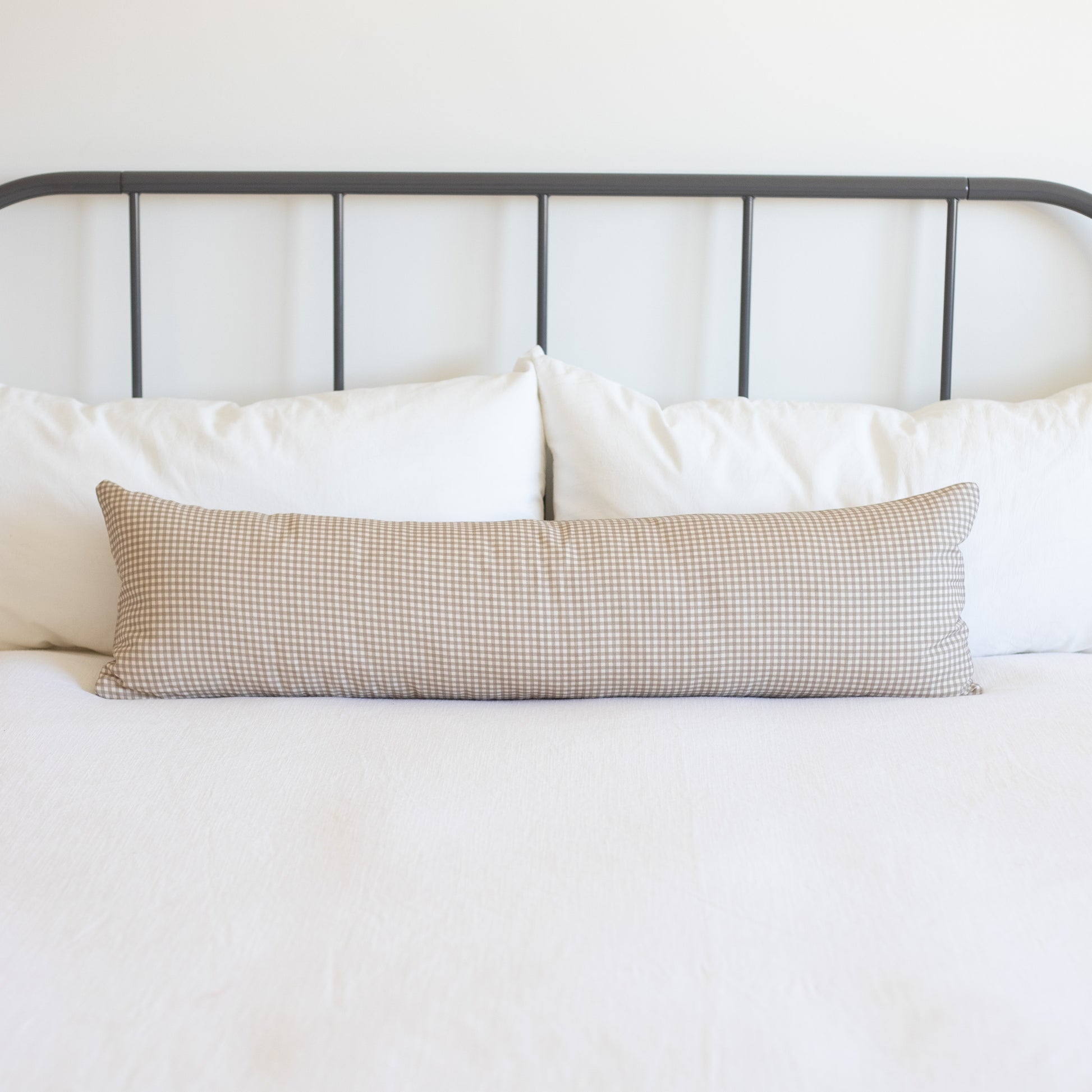 Tan and white mini gingham lumbar pillow displayed on a bed with white bedding and dark metal headboard.