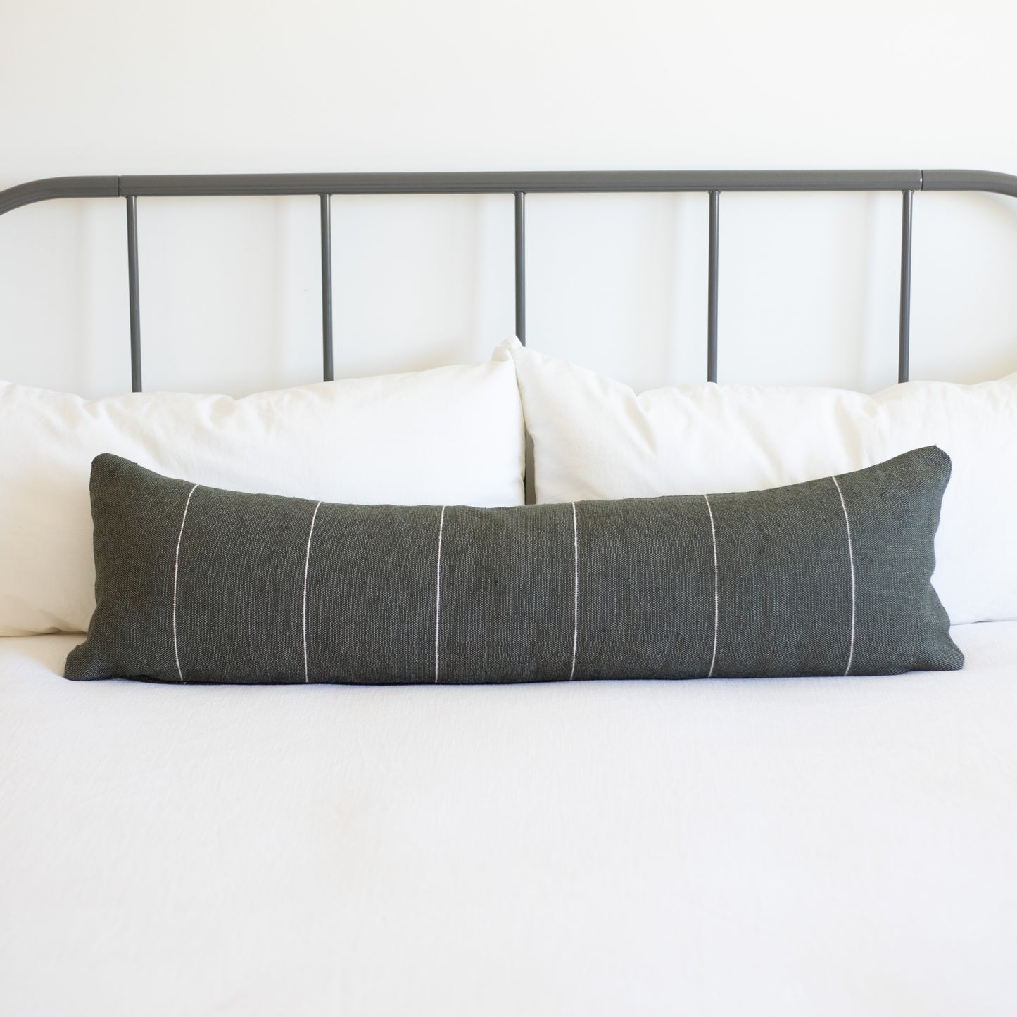 Charcoal gray lumbar pillow with white vertical stripes on bed with white pillows and metal headboard.