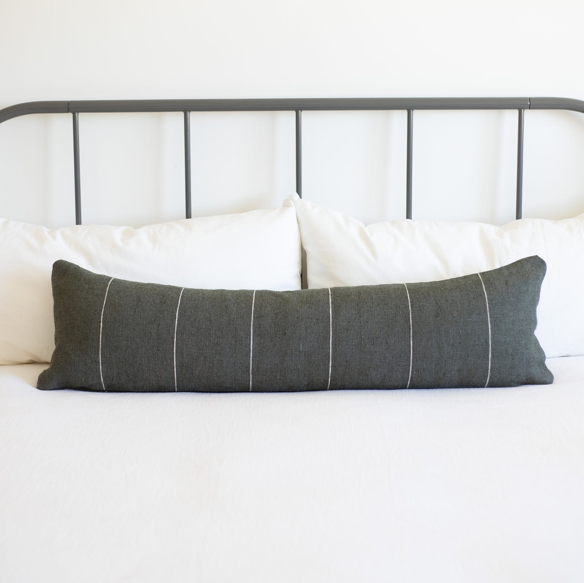 Charcoal gray lumbar pillow with white vertical stripes on bed with white pillows and metal headboard.