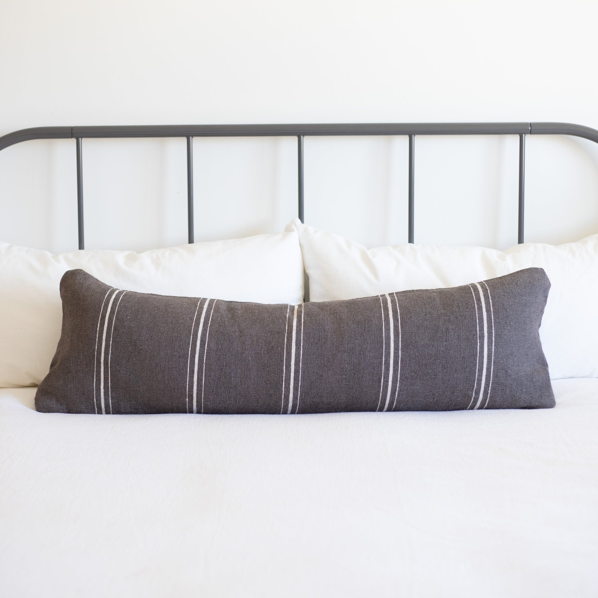 Charcoal lumbar pillow with vertical white stripes on bed with white pillows and metal headboard.