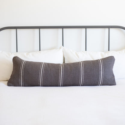Charcoal lumbar pillow with vertical white stripes on bed with white pillows and metal headboard.