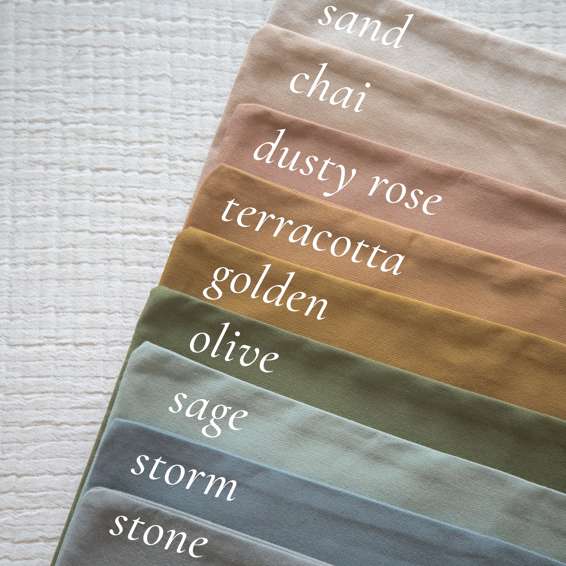 Stack of fabric swatches in neutral earth tones labeled sand, chai, dusty rose, terracotta, golden, olive, sage, storm, and s