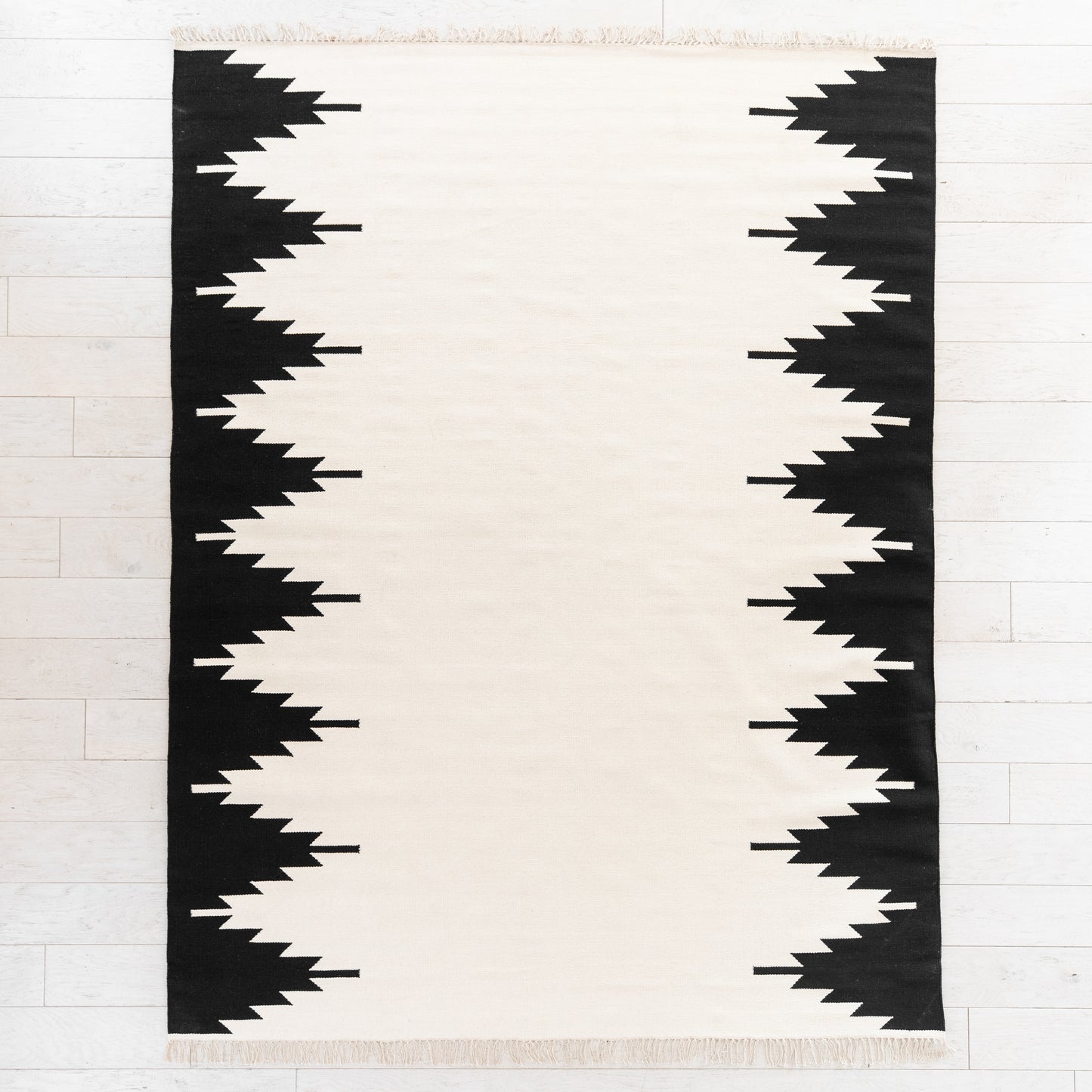 Cream and black rectangular rug with geometric jagged stripe pattern along both side edges and fringed ends.