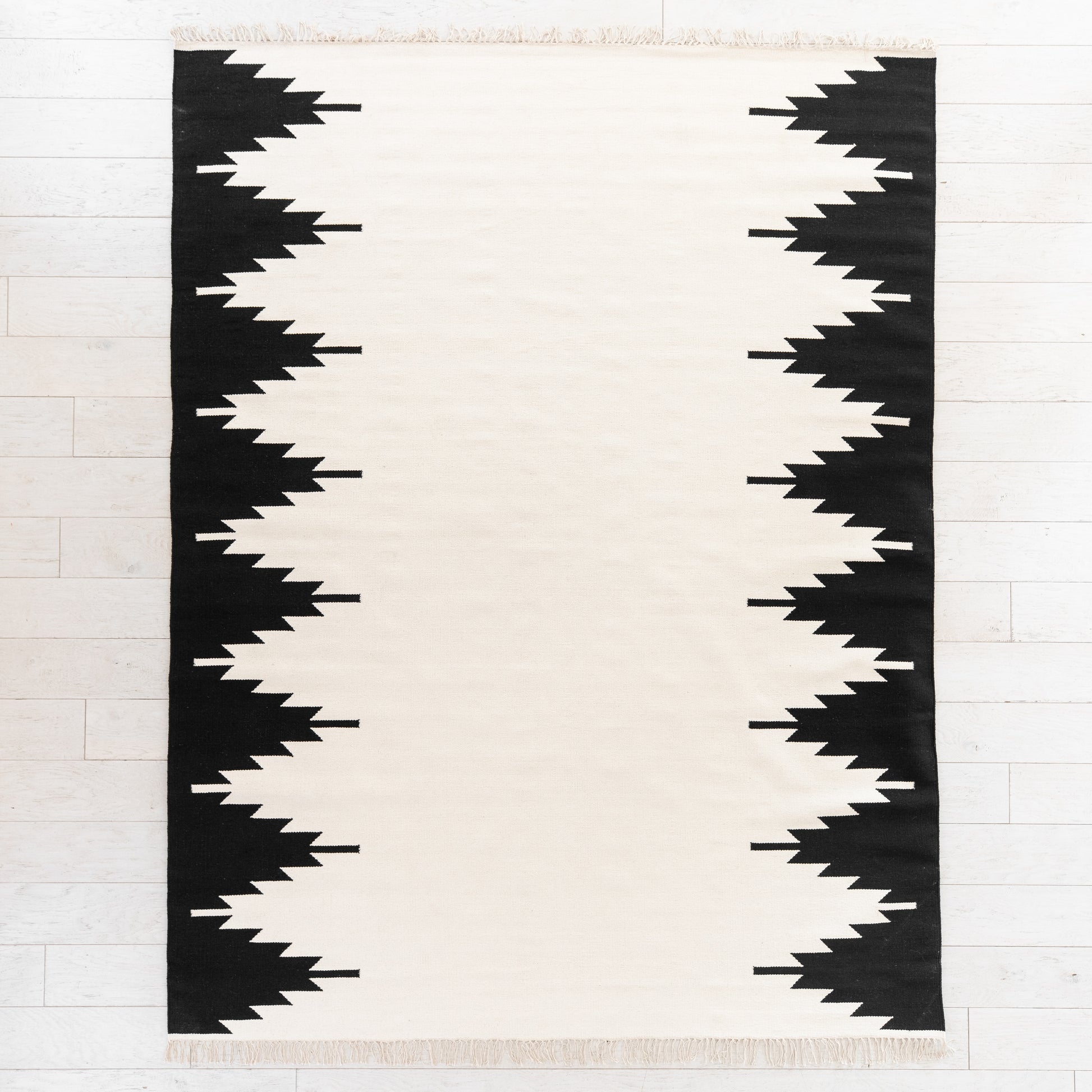 Cream and black rectangular rug with geometric jagged stripe pattern along both side edges and fringed ends.