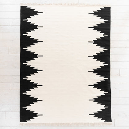 Cream and black rectangular rug with geometric jagged stripe pattern along both side edges and fringed ends.