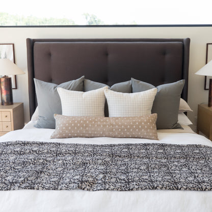 Beige lumbar pillow with white polka dot pattern displayed on bed between grid-patterned pillows.