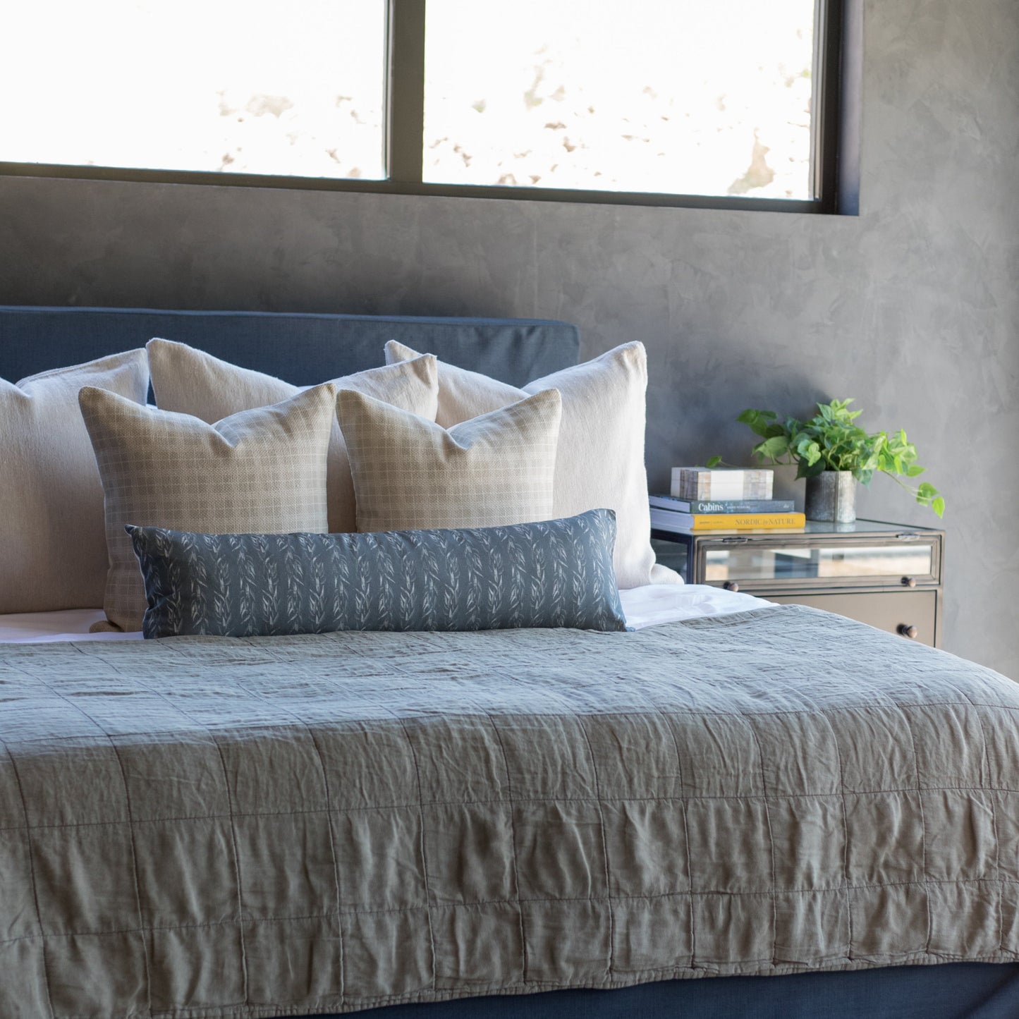 Taupe square throw pillows arranged on a bed with gray bedding and blue headboard.