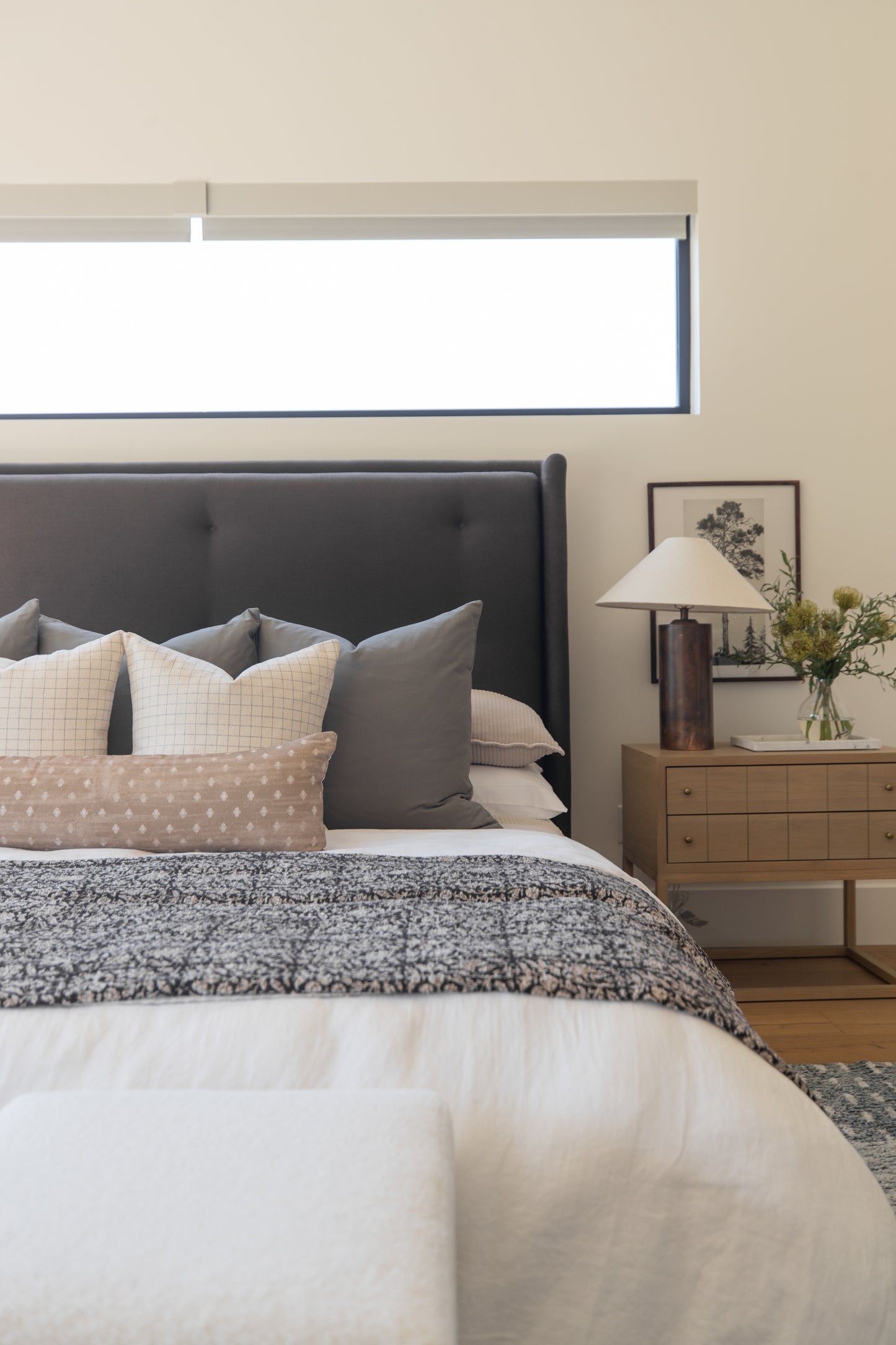 Bedroom with dark gray upholstered headboard, layered pillows in neutral tones, textured bedding, wooden nightstand with lamp