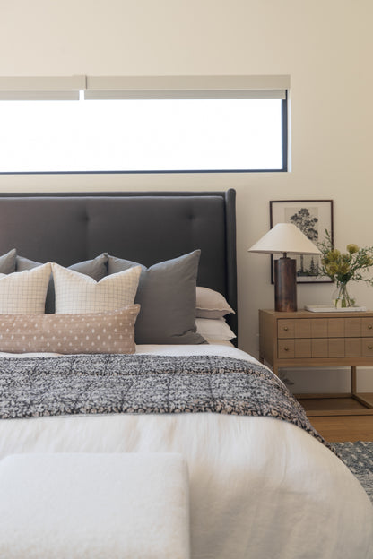 Bedroom with dark gray upholstered headboard, layered pillows in neutral tones, textured bedding, wooden nightstand with lamp