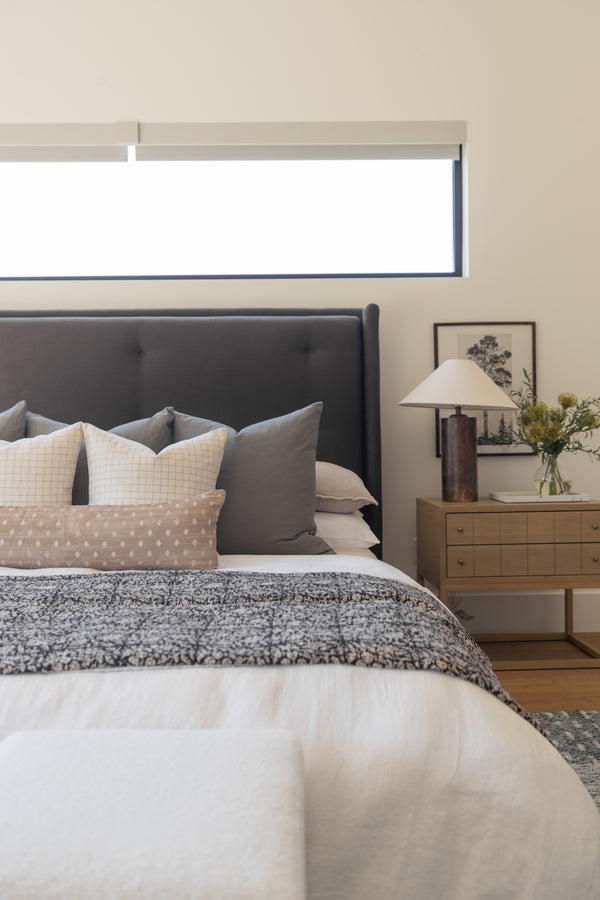 Bedroom with dark gray upholstered headboard, layered pillows in neutral tones, textured bedding, wooden nightstand with lamp