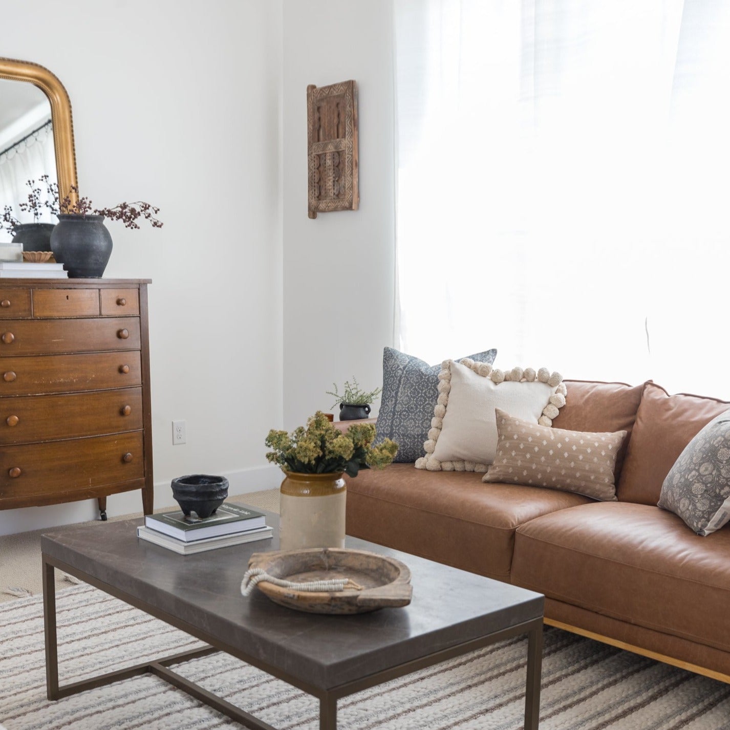 Brown leather lumbar pillow with cream pom-pom trim displayed on tan and gray sofa with decorative cushions.