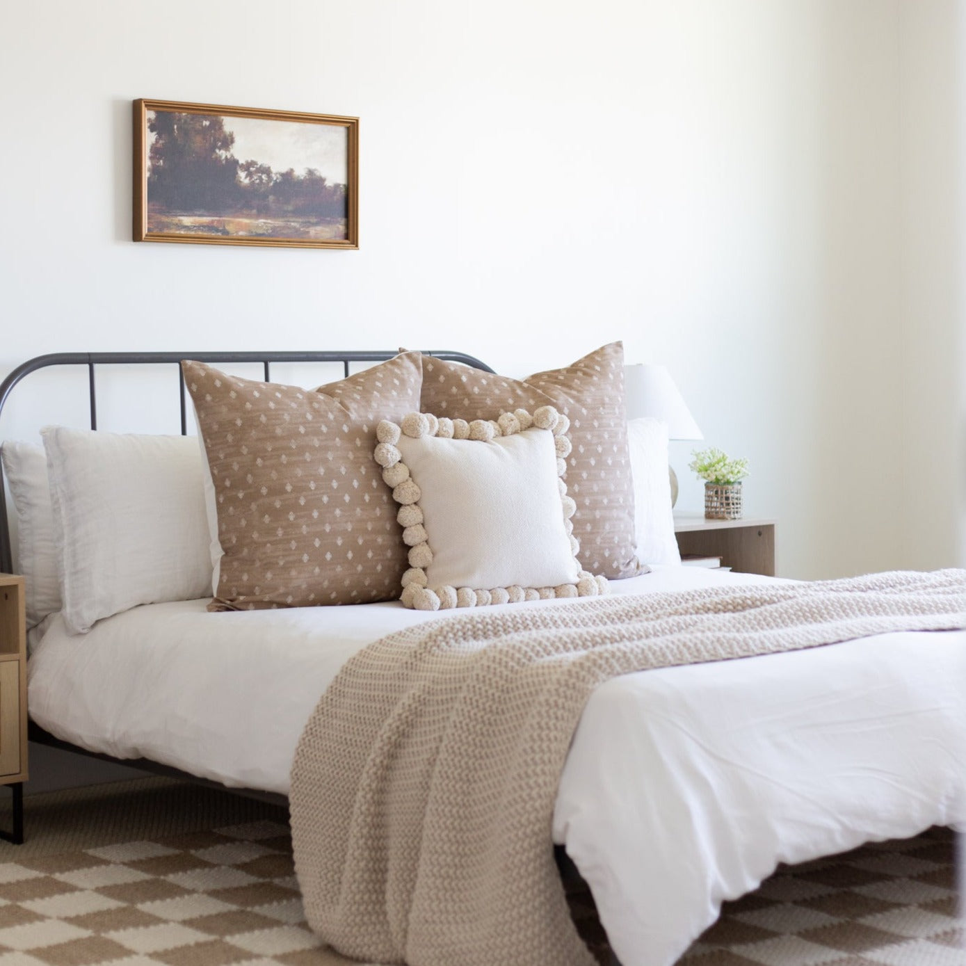 Framed landscape artwork with brown wood frame hanging above styled bed with pillows and throw blanket.