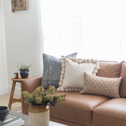 Tan leather sofa with four decorative pillows in blue patterned, cream with pom-pom trim, and tan dotted designs.