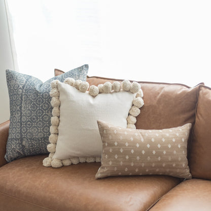 Cream lumbar pillow with pom-pom trim displayed on brown leather sofa with patterned pillows.