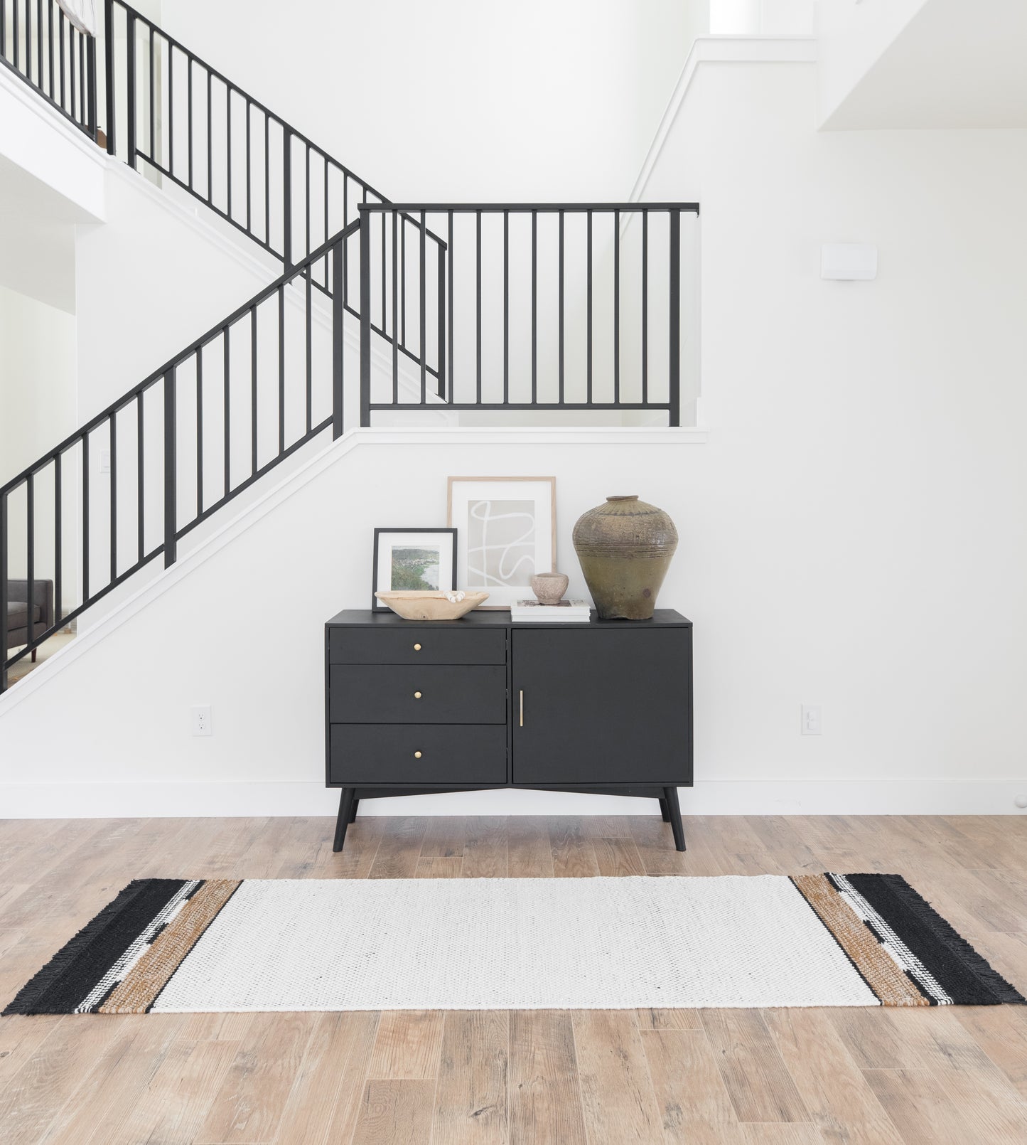 Black credenza with three drawers and cabinet door, displaying decorative items and artwork, positioned in modern entryway.