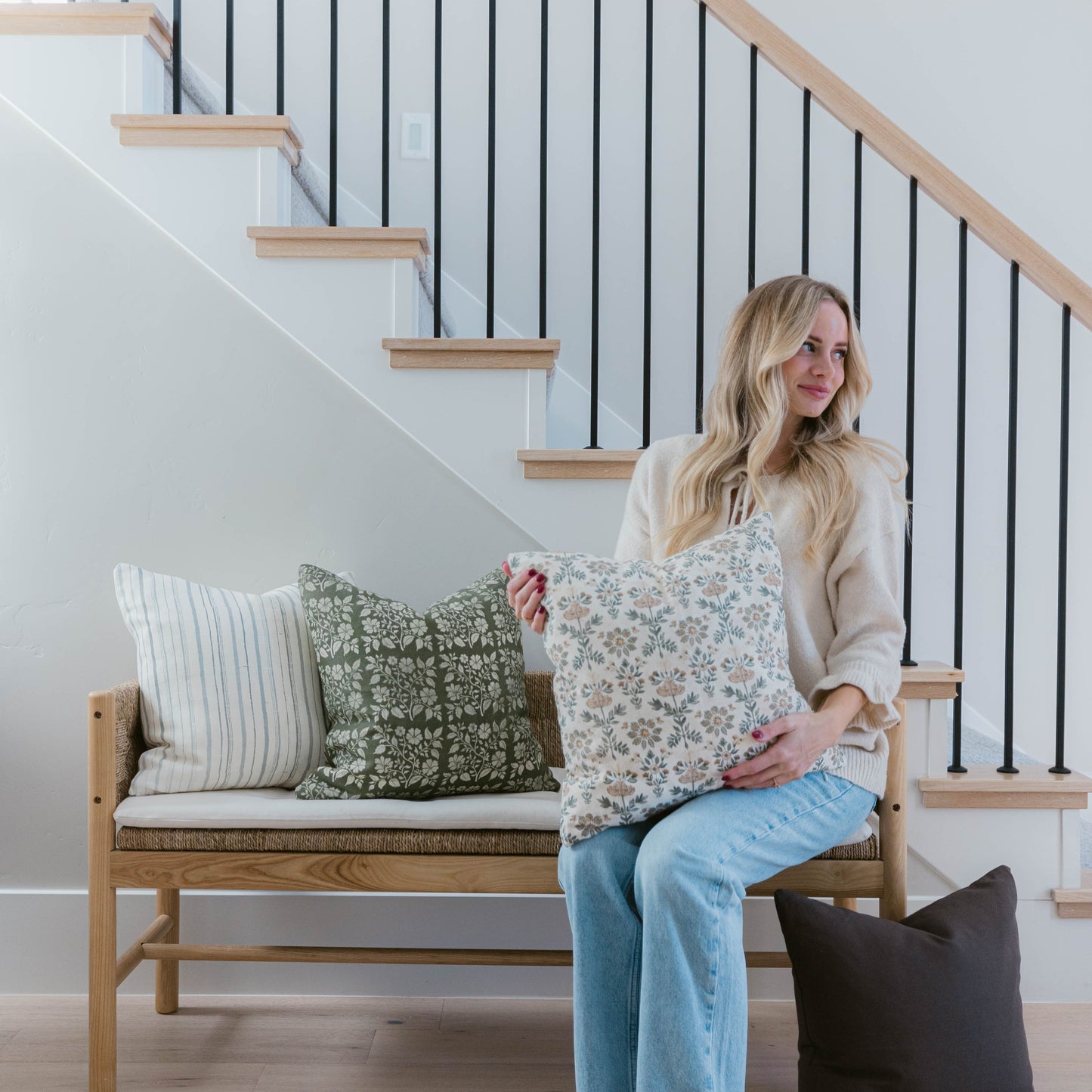 Woman sitting on wooden bench holding four decorative pillows in various patterns including stripes and florals.