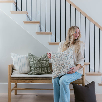 Woman sitting on wooden bench holding four decorative pillows in various patterns including stripes and florals.