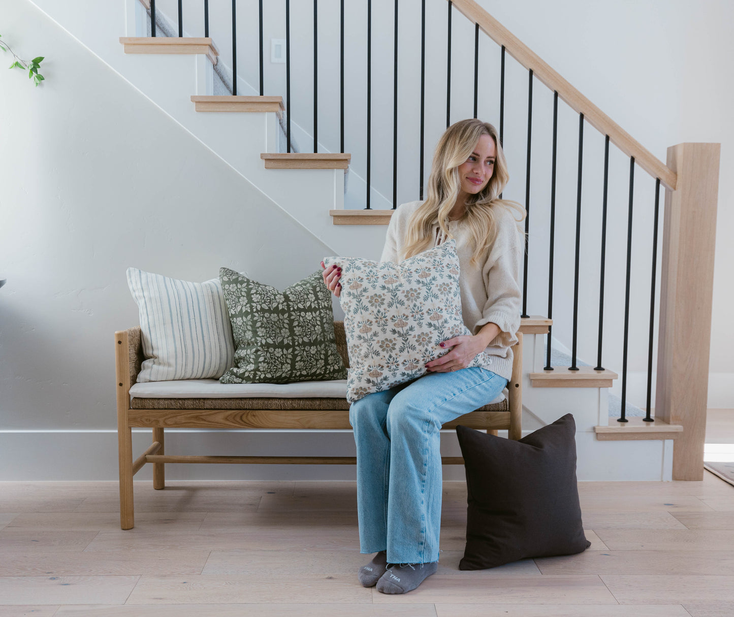 Woman sitting on wooden bench holding floral patterned throw pillow with striped and olive green pillows beside her.