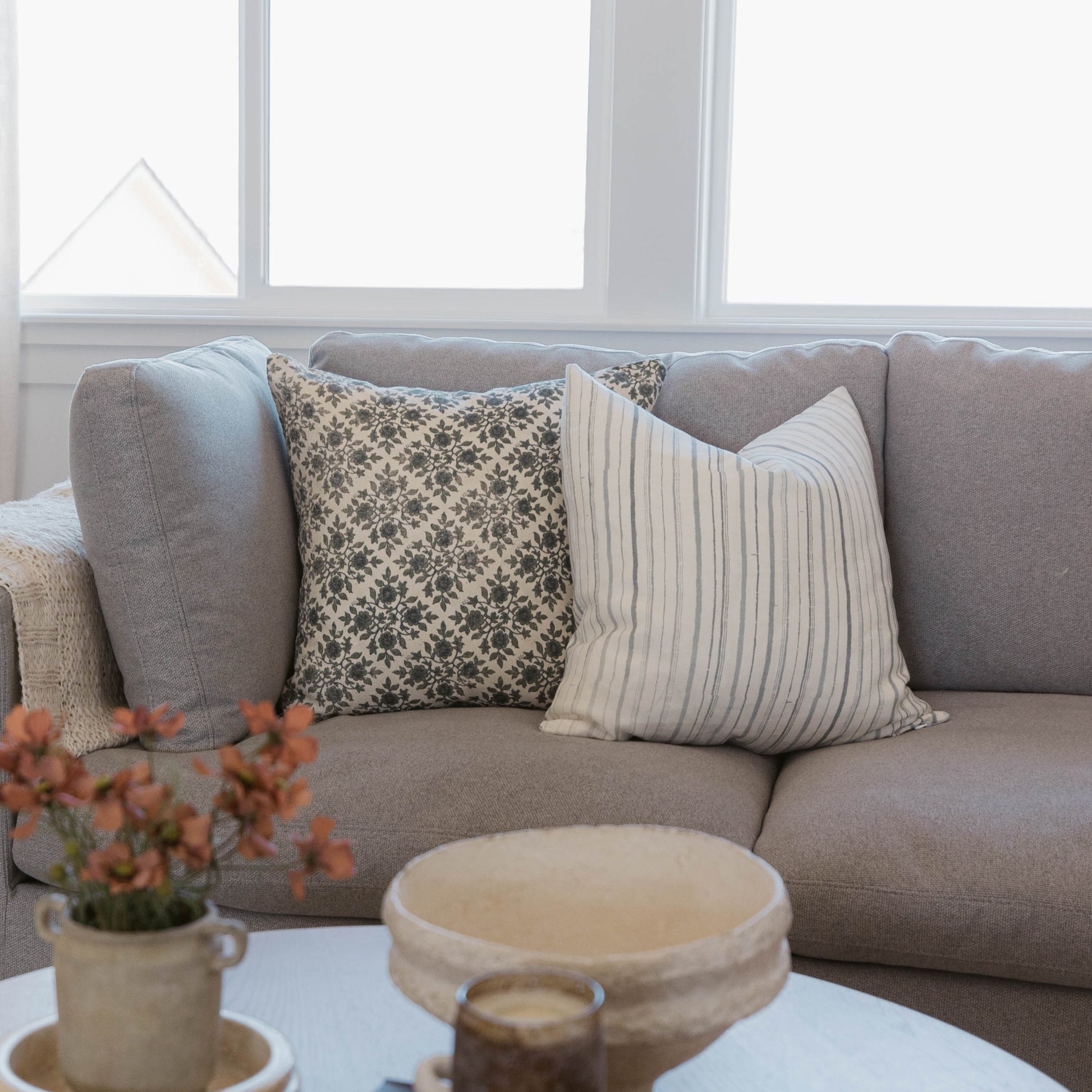 Gray linen sofa with three decorative pillows featuring floral and striped patterns, positioned near a window.