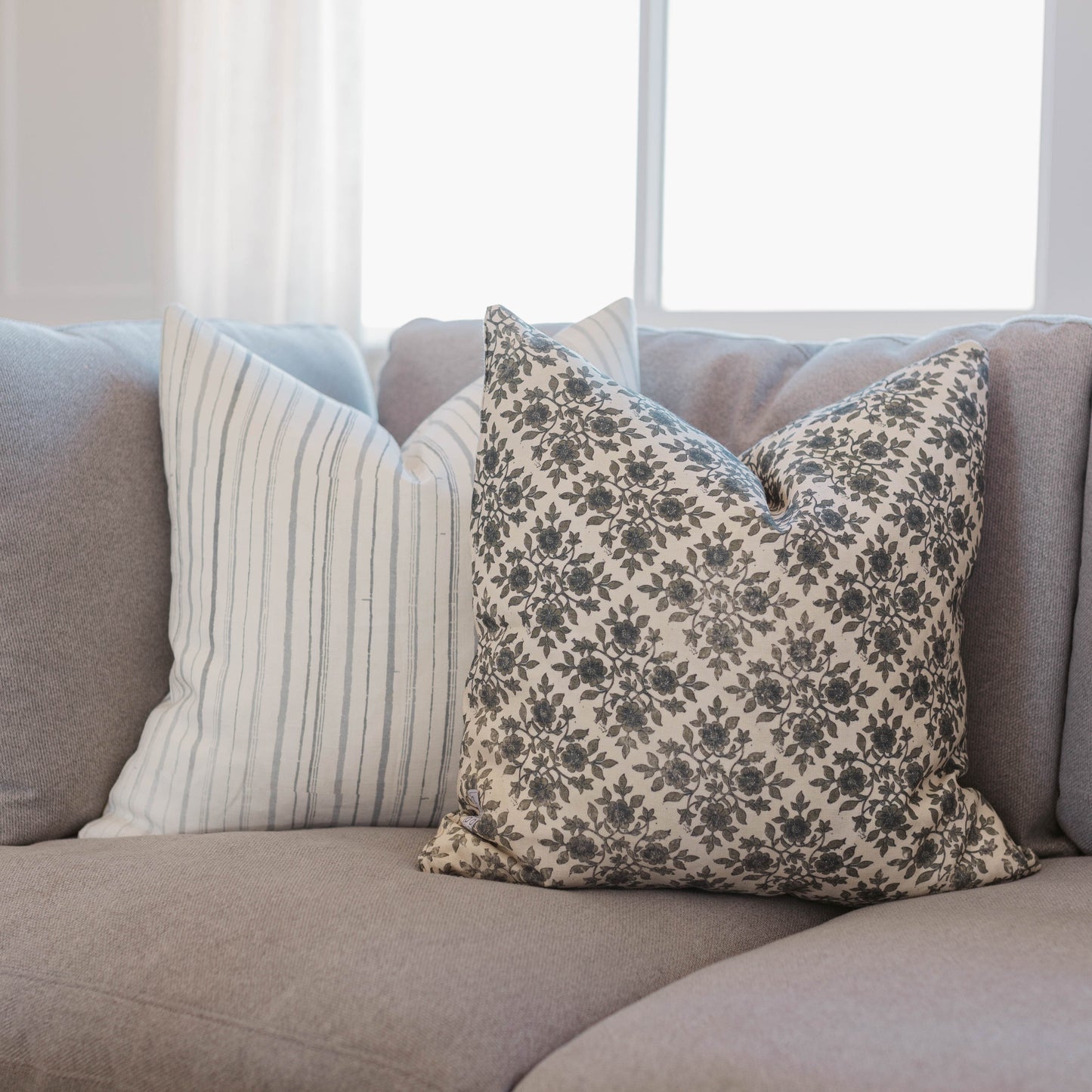 Beige linen pillow with charcoal floral damask pattern, displayed on gray sofa with striped pillow.