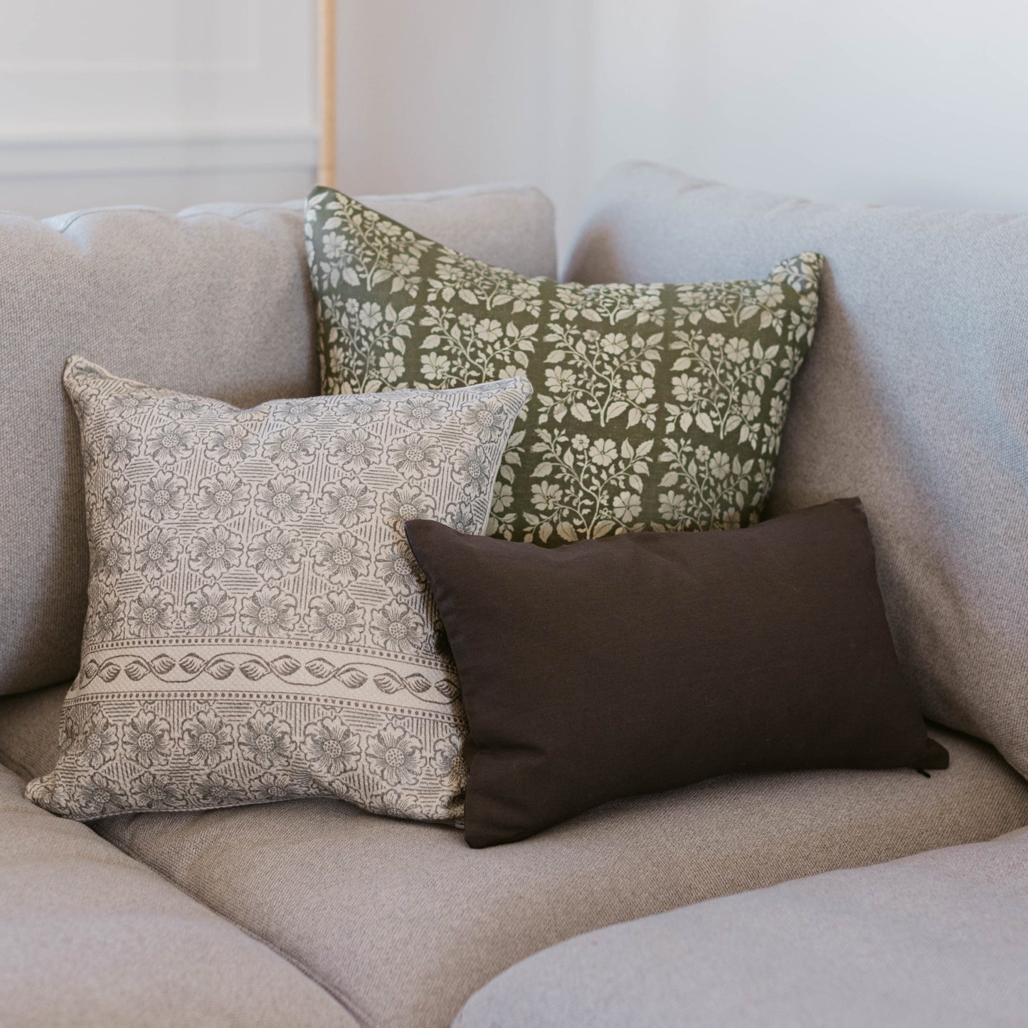 Three lumbar pillows displayed on a beige sofa: one with cream and taupe geometric pattern, one with olive green floral print