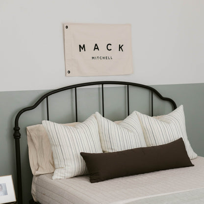 Black rectangular lumbar pillow displayed on a bed with striped pillows, shown from front view.