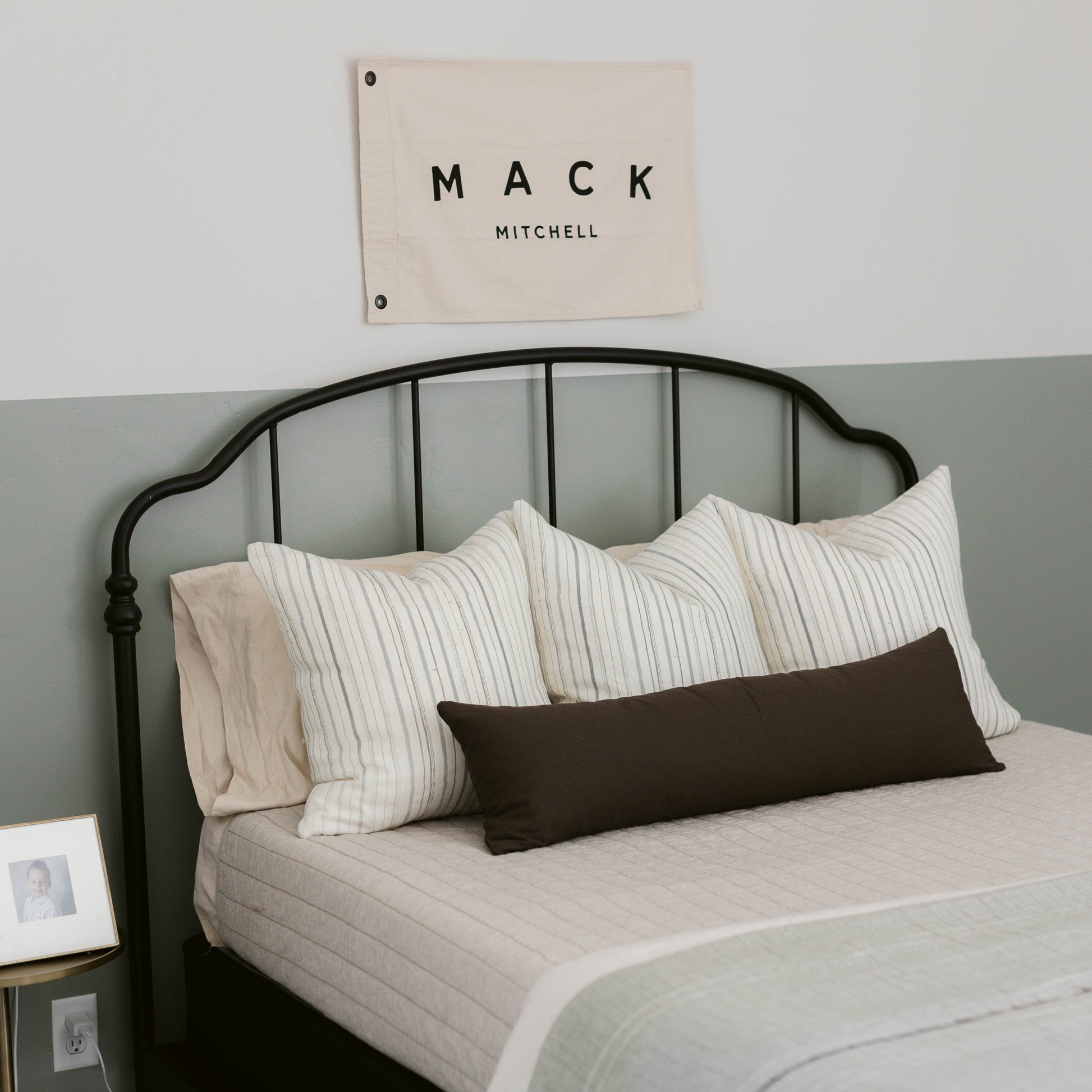 Beige linen flag with "MACK MITCHELL" text displayed above black metal bed frame with striped pillows.