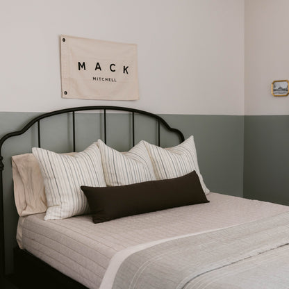 Black lumbar pillow displayed on bed with striped and solid pillows.