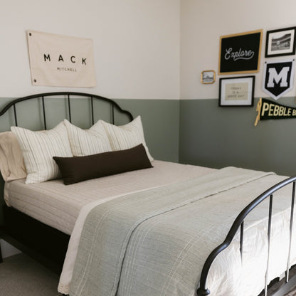 Black rectangular lumbar pillow displayed on a bed with striped pillows and a gray quilted throw.