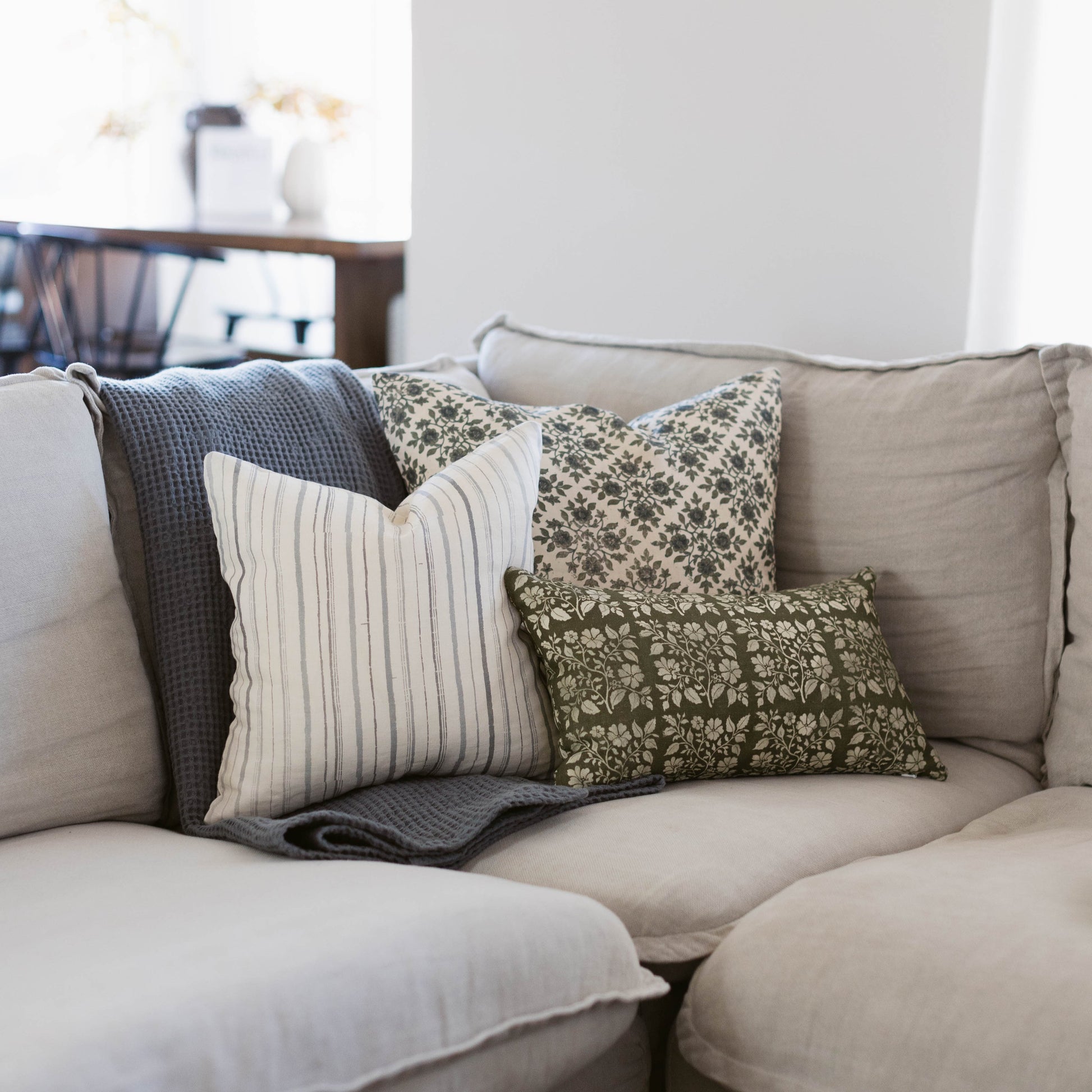 Beige linen pillow with blue piping displayed on a gray sofa with patterned and striped accent pillows.