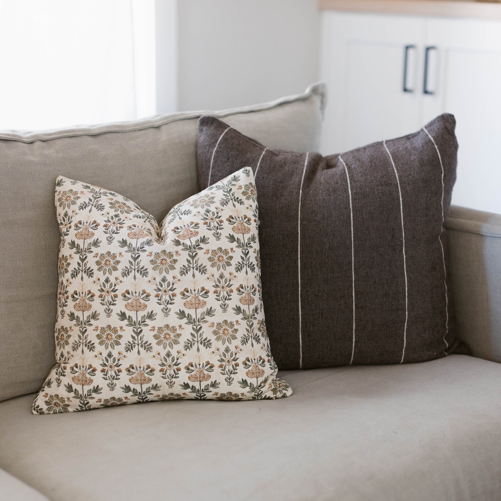 Two decorative pillows on a beige sofa: one with tan floral pattern, one with charcoal vertical stripes.