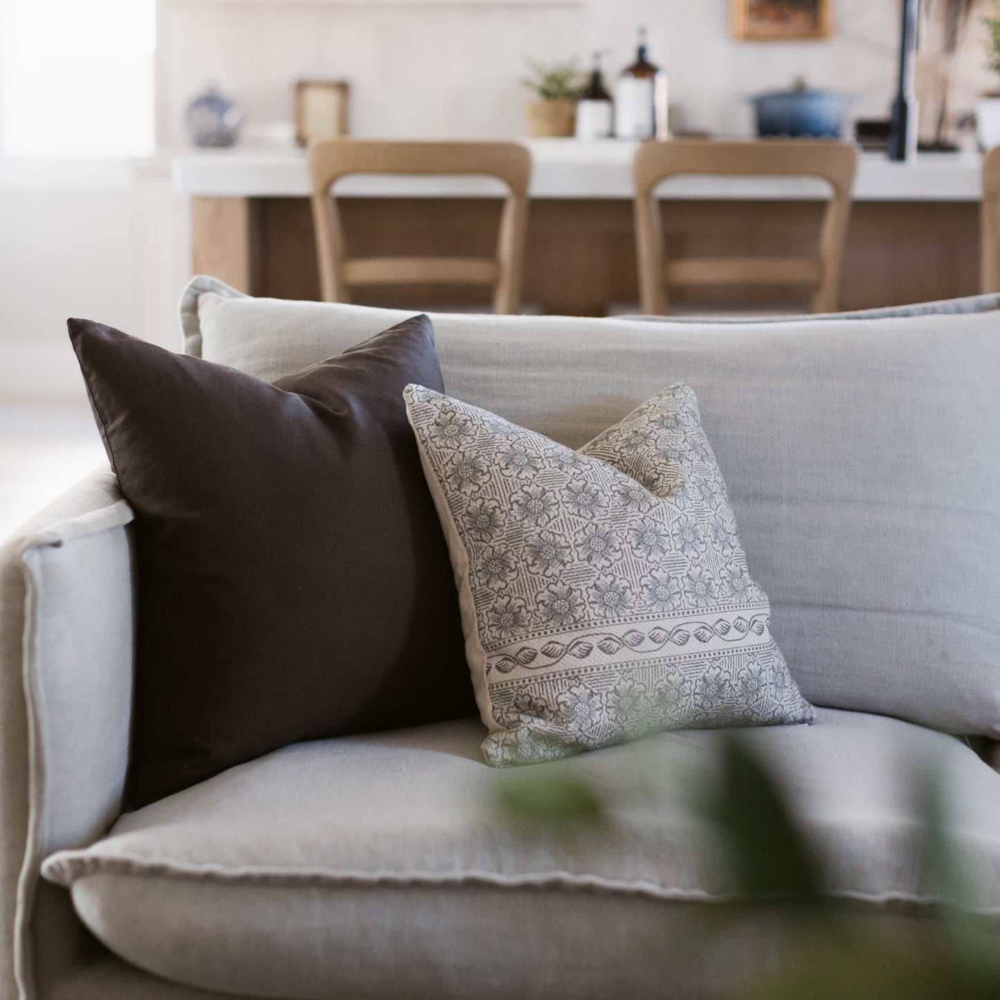 Beige patterned throw pillow with geometric design and decorative border displayed on gray sofa with black pillow.