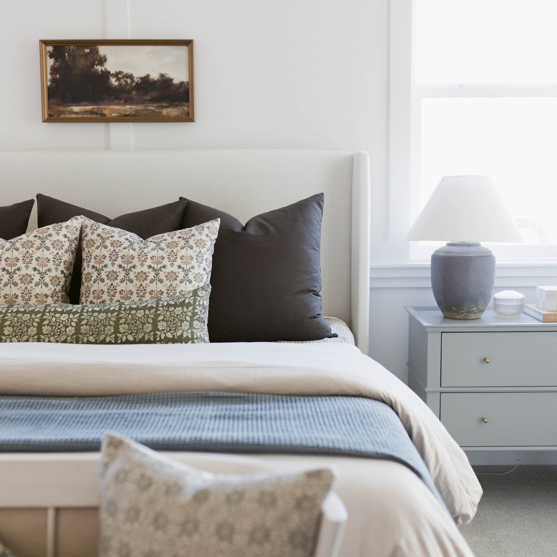 Bed styled with patterned and solid pillows, white headboard, blue throw blanket, and nightstand with lamp.