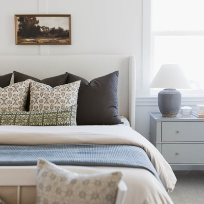 Bed styled with patterned and solid pillows, white headboard, blue throw blanket, and nightstand with lamp.
